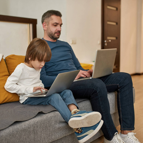 Concentrated caucasian little boy and man using and watching laptops on sofa at home. Father and son relationship and spending time together. Fatherhood and parenting. Leisure and entertainment - Photo, image
