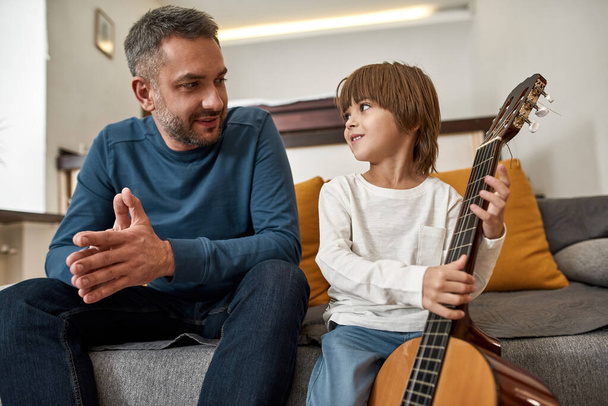 Father and little son with acoustic guitar looking at each other on sofa at home. Young smiling european family spend time together. Fatherhood and parenting. Domestic leisure, hobby and entertainment - Photo, image