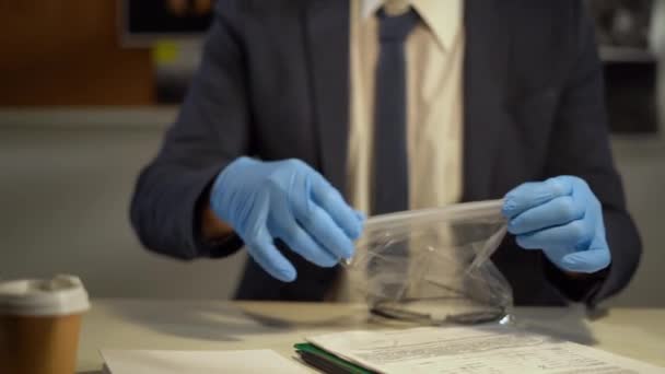mãos de um detetive de luvas azuis colocando provas em um saco no local de trabalho no escritório, um investigador examinando as provas de um caso criminal - Filmagem, Vídeo