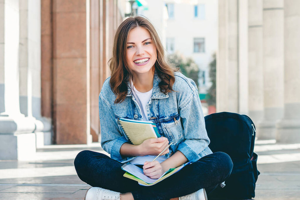 Studentessa seduta vicino all'università e sorridente. Ragazza carina studente tiene cartelle, quaderni e ride - Foto, immagini