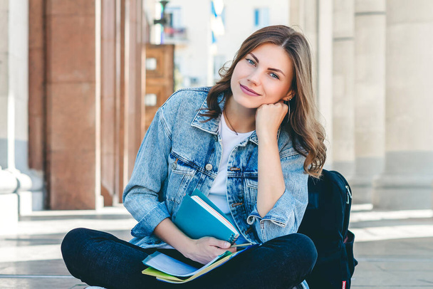 A girl sits on campus. Girl student sitting near the university and smiling. Cute girl student holds folders, notebooks. Girl teaches lessons - 写真・画像