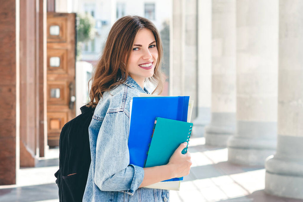 Девушка-студентка улыбается против университета. Симпатичная студентка держит в руках папки и блокноты. Обучение, концепция образования - Фото, изображение