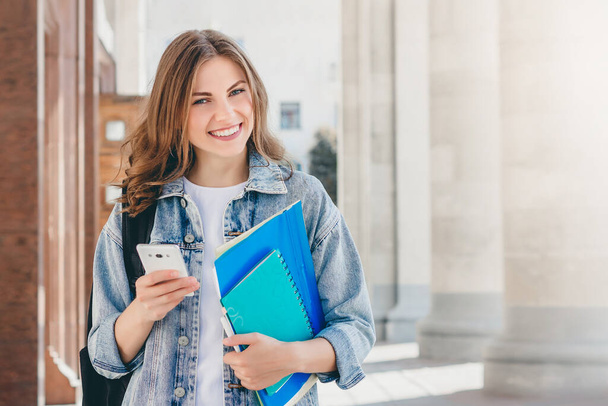 Fiatal lány diák mosolyog az egyetem ellen. Aranyos lány diák tartja mappák, notebook és mobiltelefon a kezében. Tanulás, oktatási koncepció másolási térrel - Fotó, kép