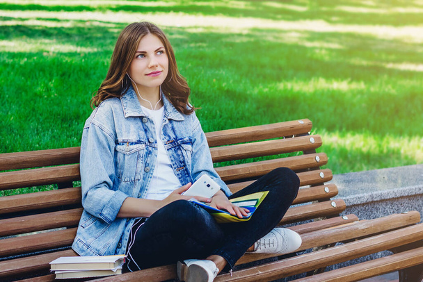 Mladá dívka student sedí na lavičce v parku a drží knihy, notebooky a mobilní telefon. Dívka poslouchá audioknihu v parku - Fotografie, Obrázek
