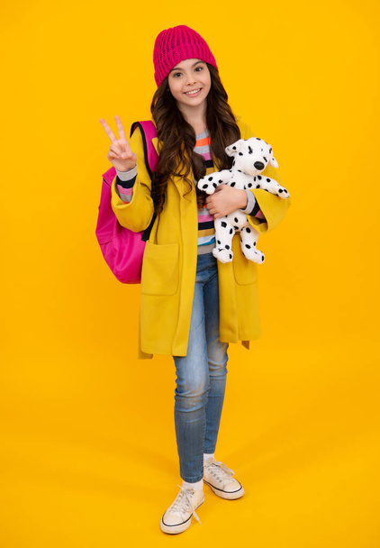 Back to school. Teenager schoolgirl in warm autumn hat hold book with toy ready to learn. School children on isolated yellow studio background - Foto, Bild