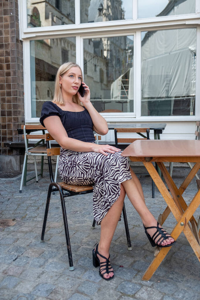 Tyylikäs nuori blondi liiketoiminnan nainen käyttää älypuhelinta istuessaan kahvilan terassilla ulkona kaupungissa. Ajatus käyttää nykyaikaista teknologiaa. Laadukas kuva - Valokuva, kuva
