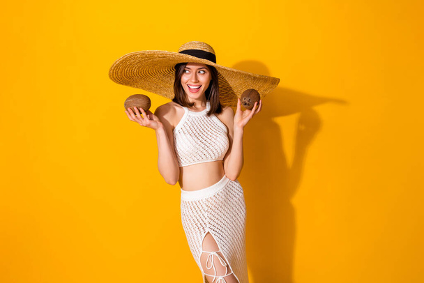Photo of positive sweet lady wear white outfit hat holding two coconuts looking empty space isolated yellow color background. - Foto, Imagen
