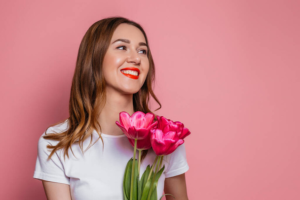Portrait of a beautiful young woman in a white t-shirt holding big bouquet of tulips isolated over pink background - Photo, Image
