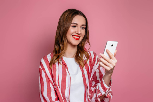 Mujer caucásica joven sonriendo y mirando a la pantalla del teléfono celular aislado en el fondo del estudio rosa. Concepto de citas online - Foto, imagen
