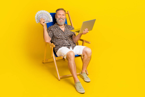 Full length photo of successful wealthy man sit lounger use netbook hold usd banknotes isolated on yellow color background. - Foto, imagen