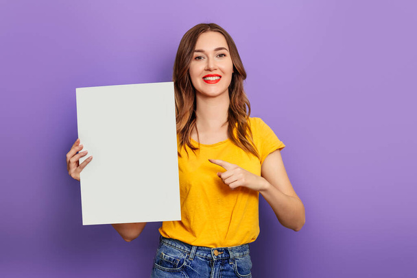 caucasian young woman holding a white poster in her hands wearing a yellow t-shirt and isolated on a lilac background. mockup - Foto, Bild