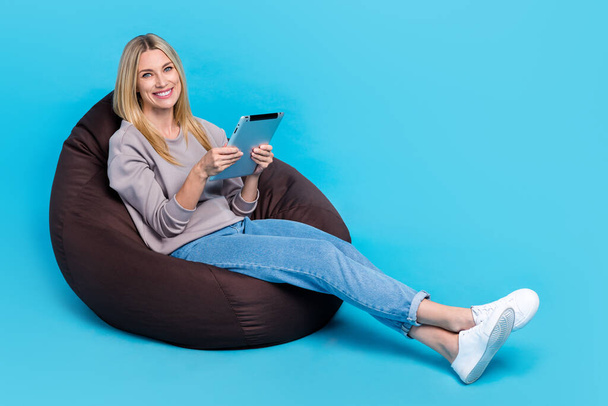 Full length photo of sweet charming lady wear grey sweatshirt typing modern gadget sitting bean bag empty space isolated blue color background. - Foto, Imagen