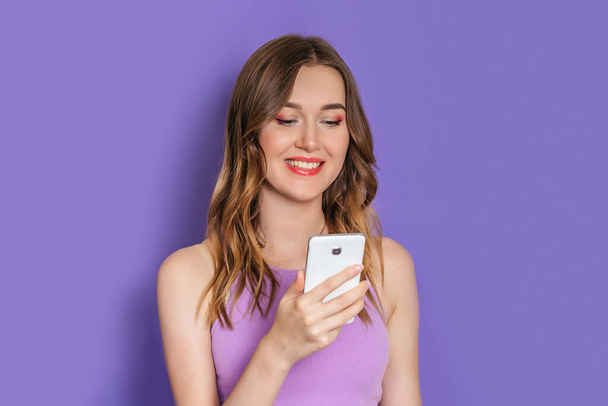 Junge kaukasische Frau mit gelocktem Haar lächelt und schaut auf den Handy-Bildschirm isoliert auf fliederfarbenem Studiohintergrund - Foto, Bild