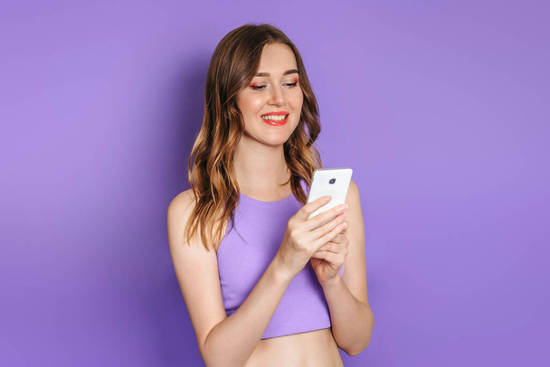 Young caucasian woman with curled hair smiling and looking at mobile phone screen isolated on lilac studio background - Photo, Image