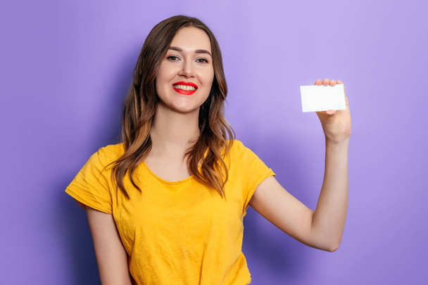 Genç beyaz bir kadın elinde bir kartvizit tutuyor ve gülümsüyor. Stüdyoda leylak bir arka planda sarı tişört giyiyor. Kredi çevrimiçi. Model - Fotoğraf, Görsel