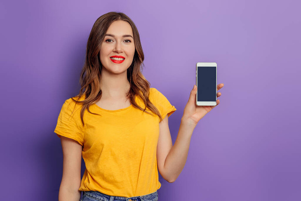 Young caucasian student girl shows a black phone screen isolated on a lilac background. Mockup - 写真・画像