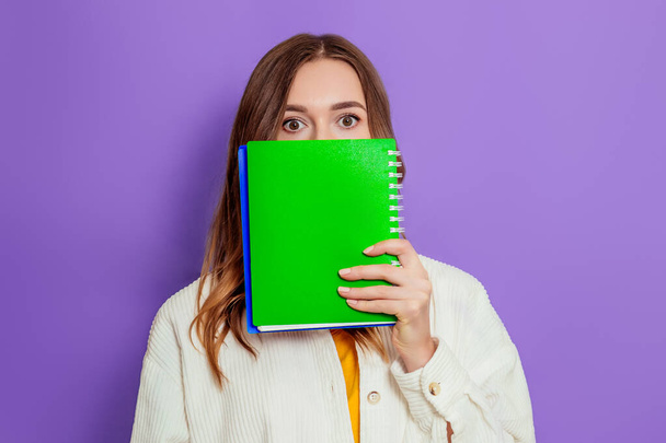 Close up portrait of a young student girl in in a beige jacket covers her face with a green exercise book isolated over purple background - Zdjęcie, obraz