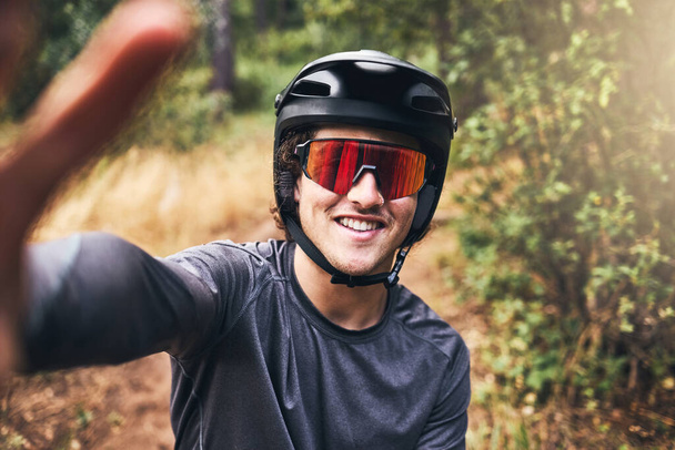 Mann macht ein Selfie beim Radfahren auf einem Naturpfad, trägt Helm und Sonnenbrille. Portrait eines Radfahrers auf einer Fahrradtour durch einen Park oder Wald, der ein Foto lächelnd und in Sicherheitsausrüstung macht. - Foto, Bild