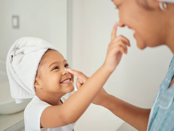 A happy Mom, smiling child using cream and teaching skincare beauty routine to girl in a bathroom. Mother helping her daughter to learn to help her skin, hair and body healthy with self care products. - Photo, image