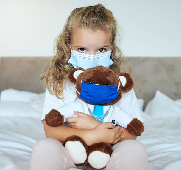 Covid, girl and teddy bear love and care for safety and awareness in pandemic quarantine at home. Portrait of a little kid playing doctor holding cute fluffy toy with mask for healthcare in pandemic. - Foto, imagen