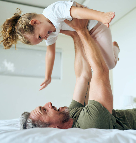 Family, dad and lifting daughter on bed while having fun and enjoying time together bonding during airplane game family activity. Playing, support and laughing man and girl child in bedroom at home. - Photo, image
