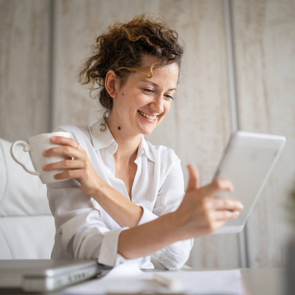 Eine junge Geschäftsfrau Kaukasierin zurück ins Büro reden online mit digitalem Tablet während der Arbeit und Kaffee trinken Unternehmer modernes Geschäftskonzept - Foto, Bild