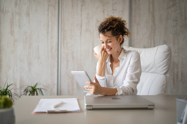 Uma jovem mulher de negócios caucasiano feminino de volta ao escritório falando on-line usando tablet digital enquanto trabalhava e bebe café empreendedor conceito de negócio moderno - Foto, Imagem