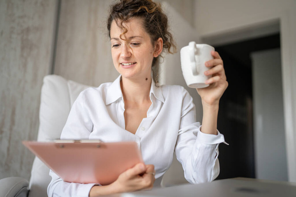 One young woman caucasian female working in the office holding clipboard and drinking coffee modern business woman entrepreneur concept - Photo, Image