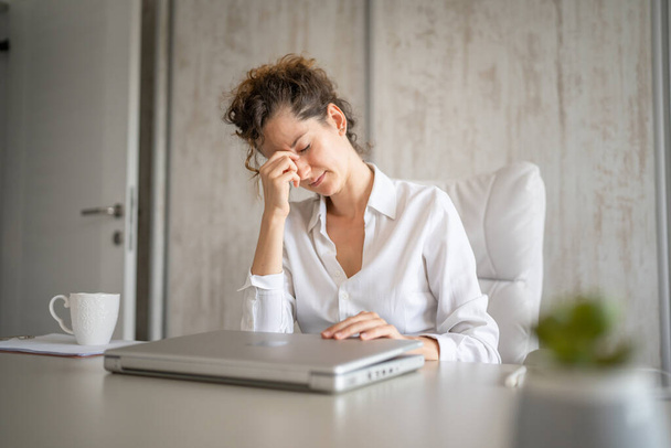 Egy nő ül az íróasztalnál a munkahelyen az irodában a székben fáradt túlterhelt és kimerült látvány miatt hosszú használata laptop számítógép valós emberek szabadúszó szemfájdalom tartsa a fejét - Fotó, kép