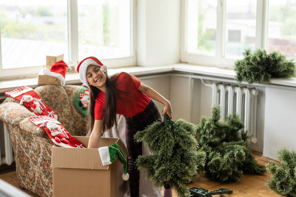 little girl makes an artificial Christmas tree wear santa hat indoors. - Zdjęcie, obraz