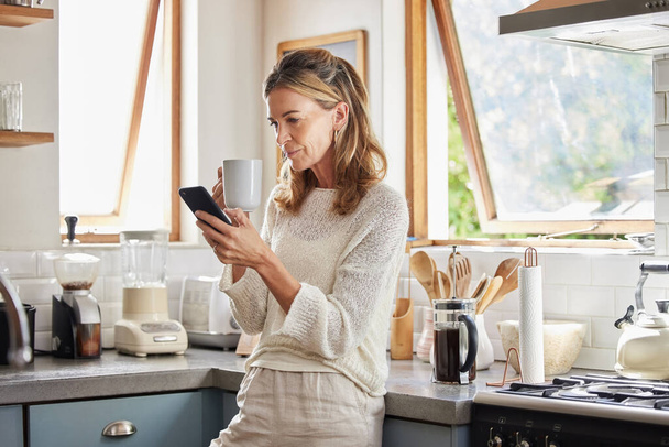 Coffee, phone and senior woman in kitchen browsing social media, text message or online app at home. Relax, tea and reading news or mobile online web surfing with female from Canada on 5g smartphone. - Фото, изображение
