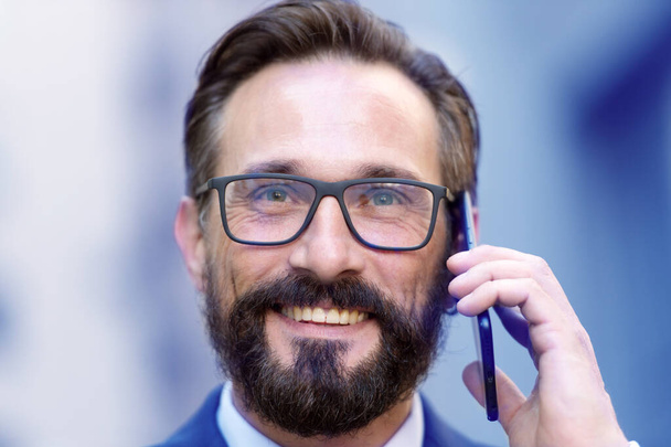 Close up of emotional bearded man with eyeswear smiling and looking happy while having phone talk. Portrait of handsome man smiling and talking on the phone - Foto, Imagem