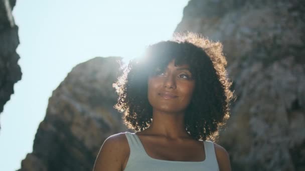 Glimlachende Afro-Amerikaanse vrouw staande rotsachtige strand verlicht fel zonlicht close-up. Portret van gekrulde jonge vrouw ontspannen na het sporten op de natuur zomerdag. Rustige sportvrouw op zoek camera buiten - Video