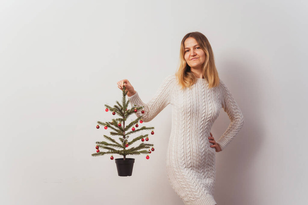 fille dans une robe tricotée avec un arbre de Noël dans ses mains sur un mur blanc. pull long tricoté à la main pour le réveillon du Nouvel An. fille avec un arbre de Noël décoré avec des jouets rouges dans la chambre - Photo, image