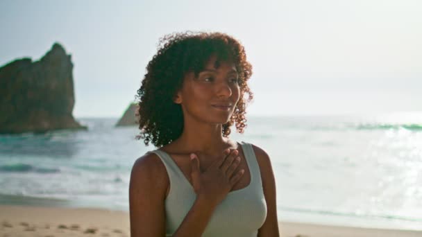 Gondtalan lány élvezni napfelkelte meditál a strandon közelről. Serene afro-amerikai nő, aki a tengerparton áll, kéz a kézben a mellkasán. Mosolygó nyugodt hölgy érzés nyugalom harmónia a természet egyedül. - Felvétel, videó