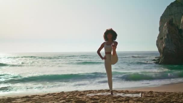 Yogi kadın güneş doğarken sahilde denge egzersizi için poz veriyor. Açık havada tek ayak üzerinde duran Afrikalı Amerikalı sporcu kız. Kum kıyısında yoga yapan esnek genç sporcu kadın.. - Video, Çekim