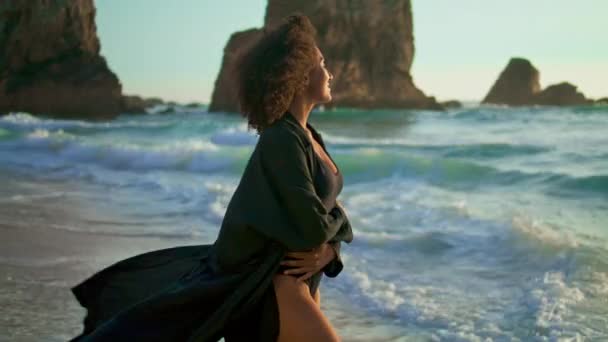 Afro-Amerikaanse ontspannen vrouw kijken naar zonsopgang staande oceaan golven op prachtig strand. Lachend aantrekkelijk meisje in sexy zwart badpak geniet van ochtendzonlicht. Kalmte harmonie op natuur concept. - Video
