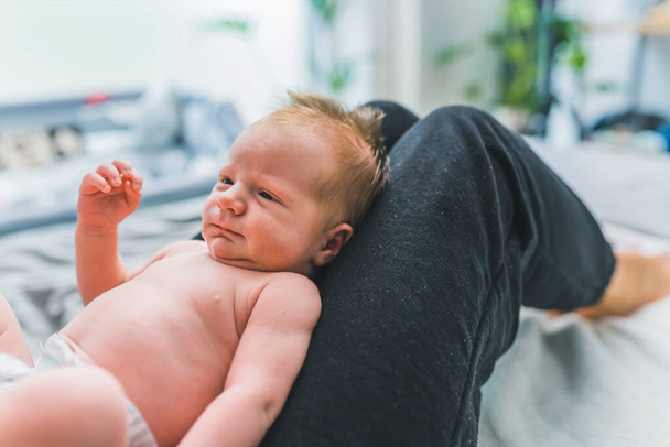 Garçon bébé caucasien aux cheveux bruns mignons posé sur les genoux de sa mère dans une couche, la regardant. Photo de haute qualité - Photo, image