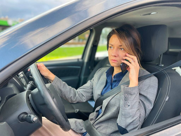 Close-up of Caucasian adult female sitting inside the car talking on cellphone with serious face expression. Businesswoman driving her automobile in city while calling on smartphone. Work life - Фото, изображение