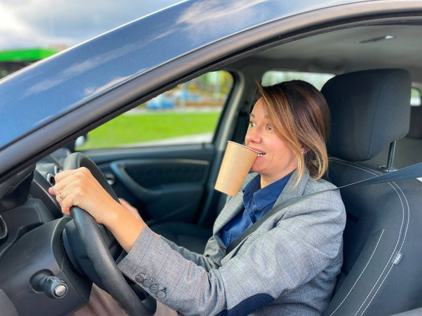 widok z boku dość zabawne businesswoman jazdy samochodem w mieście z ruchu trzymając filiżankę kawy w ustach z dużymi oczami patrząc na drodze z zaskoczoną twarzą. Biała kobieta prowadzi auto z kawą - Zdjęcie, obraz