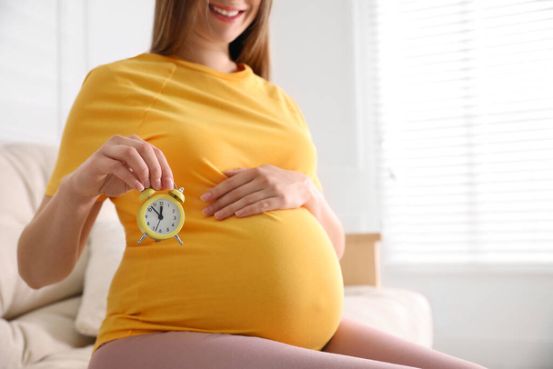 Nuori raskaana oleva nainen pitelee herätyskelloa vatsansa lähellä kotona, lähikuva. Synnytysaika - Valokuva, kuva