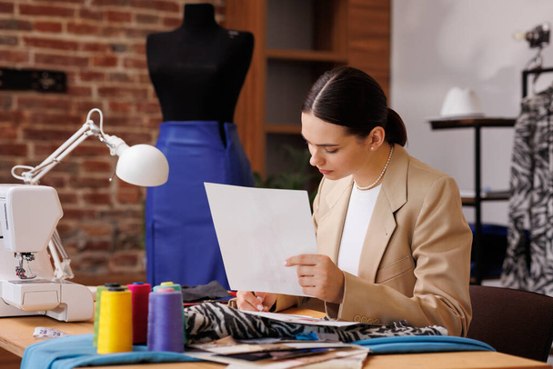 A young girl is a fashion designer, draws a sketch against the background of a mannequin and a sewing machine, in her cozy office. - 写真・画像