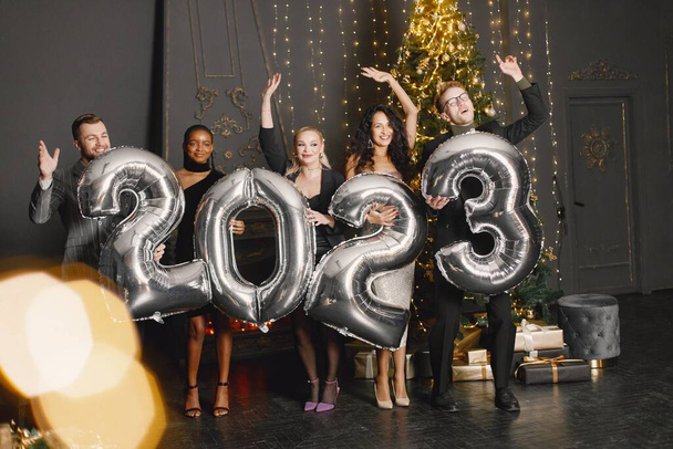 Young women with a hairstyle in an elegant outfits and man in costumes holding a ballons in shape of numbers 2023. Women standing in the decorated interior of the house during the Christmas holiday - Foto, Imagem