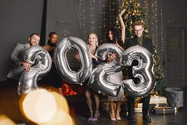 Young women with a hairstyle in an elegant outfits and man in costumes holding a ballons in shape of numbers 2023. Women standing in the decorated interior of the house during the Christmas holiday - Foto, Imagen
