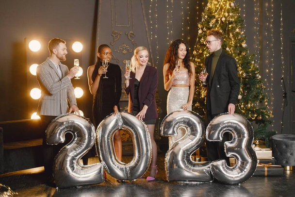 Young women with a hairstyle in an elegant outfits and man in costumes holding a ballons in shape of numbers 2023. Women standing in the decorated interior of the house during the Christmas holiday - Foto, Imagen