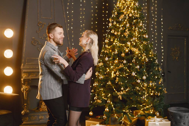 クリスマスツリーによって装飾されたリビングルームでロマンチックなカップルダンス。新年のパーティーを祝うカップル。エレガントな衣装と女性の黒のドレスを着た男. - 写真・画像