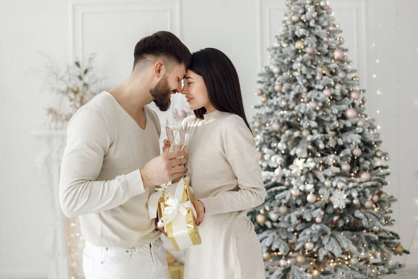 Romantic couple celebrating Christmas near Christmas tree. Brunette woman and man wearing white clothes. Romantic couple holding a gifts and glasses of champagne. - 写真・画像