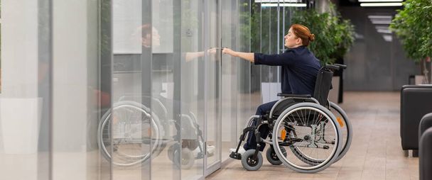 Red-haired caucasian woman in a wheelchair trying to open the door in the office - 写真・画像
