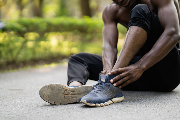 Afrykański sportowiec biegacz siedzi na torze joggingowym używając ręki trzymając kostkę i czując ból w kostce, przestrzeń kopiowania. Koncepcja urazu sportowego - Zdjęcie, obraz