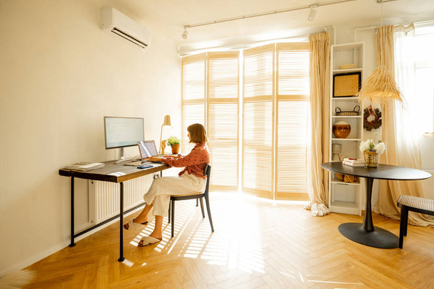 La donna lavora al computer mentre si siede vicino a un accogliente posto di lavoro nel soleggiato soggiorno di casa. Concetto di lavoro a distanza dall'ufficio di casa - Foto, immagini
